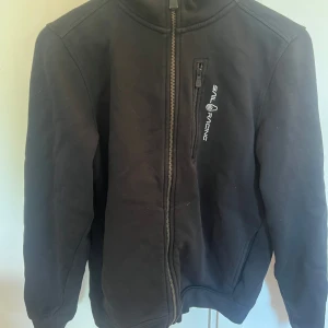 Svart Sail Racing jacka med huva - Svart jacka från Sail Racing med huva och dragkedja framtill. Jackan har en bröstficka med dragkedja och logga på bröstet samt på huvan. Justerbar snörning i huvan och ribbade muddar. Perfekt för dig som gillar stilren och sportig look.