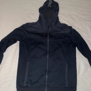 Mörkblå zip kofta Hugo Boss strl L - Snygg mörkblå zip kofta från Hugo Boss med broderad logga på bröstet. Tröjan har dragkedja, huva med snörning och ribbade muddar. Perfekt för en chill och stilren look. Materialet känns mjukt och bekvämt, troligen bomullsmix. Jätte lite sliten nere vid handleden enkelt att sy men man tänker inte alls på det :) väldigt fin kollektion som ej finns nå mer på marknaden.