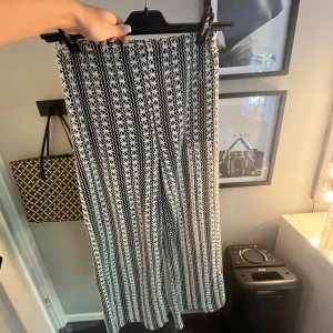HM stackade byxor  - Säljer ett par svarta och vita byxor med blommigt mönster och elastisk midja. Byxorna har ett luftigt, wide fit och är perfekta för dig som gillar statement-plagg. Materialet är stickat och mönstret är riktigt unikt med broderade blommor.