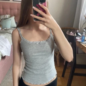 Grå ribbad linnetopp med volangkant - Supersöt grå ribbad linnetopp med smala axelband och volangdetaljer längs kanterna. Toppen har en liten rosett framtill och vågig nederkant. Perfekt för en chill och trendig look.