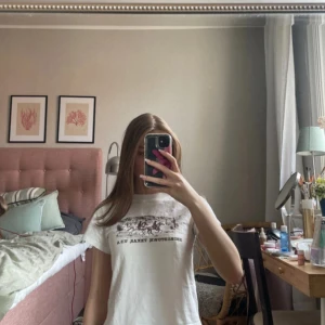 Vit t-shirt Georgetown Texas print - Vit t-shirt med brunt tryck framtill som föreställer cowboys och texten 'Georgetown, Texas, U.S.A'. Modellen är croppad med rund hals och korta ärmar. Perfekt för dig som gillar en chill och avslappnad stil.