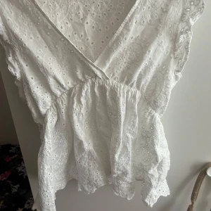 Vit broderad blus med volang - Superfin vit blus i bomull med broderade detaljer och volangkant. Omlottmodell med v-ringning och lätt rynkad midja som ger en söt och luftig känsla. Perfekt för varma dagar när du vill ha något romantiskt och fräscht.