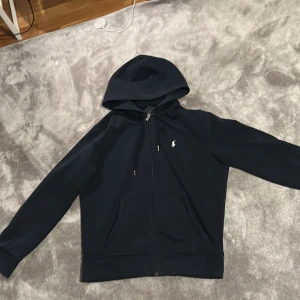 Svart hoodie från Polo Ralph Lauren - Snygg mörkblå hoodie från Polo Ralph Lauren med dragkedja och klassisk logga på bröstet. Tröjan har huva med snörning och två fickor framtill. Inga defekter, och i ett mycket bra skick 8/10. 