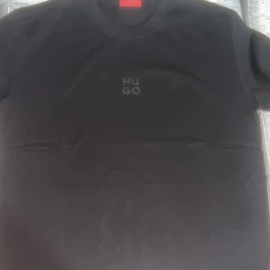 Svart t-shirt från Hugo Boss - Svart t-shirt från Hugo Boss med diskret HUGO-tryck på bröstet. Klassisk rund halsringning och korta ärmar. Perfekt basic-plagg med stilren vibe och mjuk känsla.