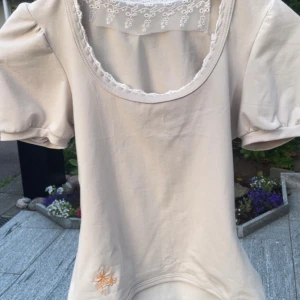 Beige blus med spets och puffärm - Supersöt beige blus med puffiga korta ärmar och rundad halsringning dekorerad med spets. Nedtill finns en liten broderad detalj i orange/korall 🧡hemmasydd av mig, sitter figurnära o passar XS eller liten S
