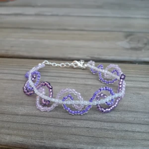 Lila och vit pärlarmband med swirl - Handgjort armband med små pärlor i lila, vitt och ljuslila. Pärlorna är trädda i ett spiral/vågor mönster som ger en cool och lekfull look. Armbandet har justerbar silverfärgad kedja och karbinhake. Perfekt för dig som vill sticka ut med färg och mönster.