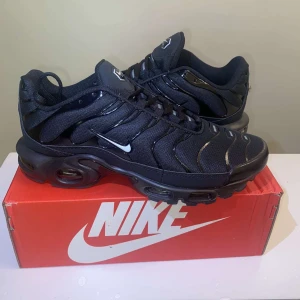 Svarta Nike sneakers med luftdämpning Nike tn  - Snygga svarta Nike sneakers med ikonisk vågig design och synlig luftdämpning i sulan. Skorna har mesh och syntetmaterial, svarta snören och en liten vit swoosh på sidan. Perfekta för dig som gillar streetstyle och vill ha bekväma skor med cool look.