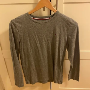 Grå långärmad tröja Tommy Hilfiger - Snygg grå långärmad tröja från Tommy Hilfiger med diskret logga på bröstet. Klassisk rund halsringning med röd, vit och marinblå detalj på insidan av kragen. Perfekt basic-plagg för en clean och stilren look.