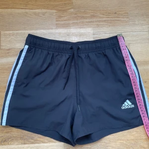 Svarta Adidas shorts med snörning S - Svarta sportiga shorts från Adidas med vita ränder längs sidorna och klassisk logga framtill. De har elastisk midja med snörning och är tillverkade i lätt polyester med meshfoder. Perfekta för träning eller chill dagar på sommaren.