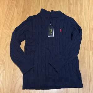 Marinblå stickad tröja Polo Ralph Lauren - Snygg marinblå stickad tröja från Polo Ralph Lauren med half zip och klassisk röd logga på bröstet. Tröjan har kabelstickat mönster, hög krage och långa ärmar. Perfekt för dig som gillar stilren och tidlös design. 