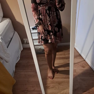 Svart blommig omlottklänning - Supersöt svart kortklänning med stora rosa blommor och v-ringning. Klänningen har omlottmodell, markerad midja och långa ärmar med lätt puff. Perfekt för dig som gillar romantiska vibes och flowy kjol.