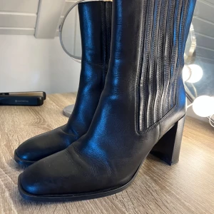 Svarta skinnstövletter med klack - Snygga svarta stövletter i skinn med markerad fyrkantig tå och bred klack. Sidan har dekorativa vertikala sömmar som ger en cool touch. Perfekta för dig som gillar stilrena boots med lite edge.