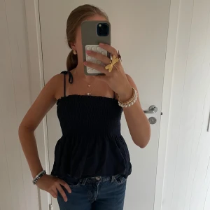 Mörkblå smockad linnetopp med knyt - Supersöt mörkblå linnetopp med smockad överdel och volang nertill. Axelbanden knyts på axlarna för en personlig touch. Perfekt till jeans eller kjol för en snygg look. 