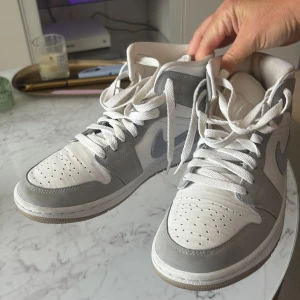 Nike Air Jordan 1 Mid vit/grå - Nike Air Jordan 1 Mid i fräscha vita och grå toner. Klassisk high-top siluett med snörning, perforerad tå och ikonisk Air Jordan-logga på sidan. Ovandel i skinn och mocka med grå Swoosh. Perfekta sneakers för dig som gillar streetstyle.