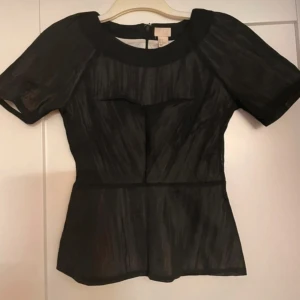 Svart blus med dragkedja från H&M - Snygg svart blus från H&M med korta ärmar och markerad midja. Blusen har en diskret struktur i tyget, rund halsringning och dragkedja bak. Tunt, lätt material med innerfoder för extra komfort. Perfekt för dig som gillar stilrena och trendiga plagg.