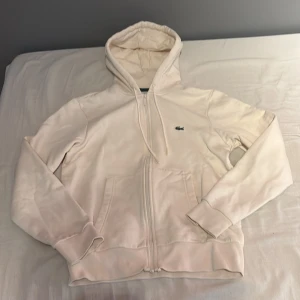 Vit hoodie från Lacoste med dragkedja - Säljer en stilren vit hoodie från Lacoste i klassisk passform. Tröjan har huva med snörning, dragkedja framtill och två fickor. Ikoniska krokodilloggan broderad på bröstet. Perfekt för dig som gillar clean och enkel stil.Storlek xs men sitter som en S