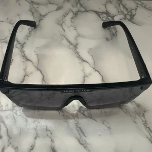 Svarta solglasögon - Snygga svarta solglasögon  med breda skalmar och guldigt LV-logo på sidan. Glasen är mörka och har en rak, futuristisk form som ger en riktigt cool vibe. Perfekta för dig som vill sticka ut och köra på en lyxig streetstyle. Vid snabb affär kan priset diskuteras.