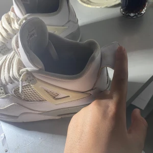 Nike Air Jordan 4 Retro vita sneakers - Säljer ett par Nike Air Jordan 4 Retro sneakers i vitt skinn med meshpaneler och grå detaljer. Skorna har den klassiska Jumpman-loggan i rött på plösen och bak på hälen. Kraftiga vita snören och ikoniska plastdetaljer på sidorna. Perfekt för dig som gillar streetwear äkta.