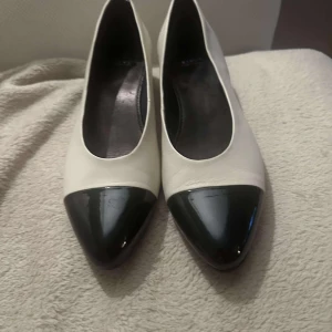 Svartvita pumps med låg klack - Stilrena pumps i vitt skinn med svart, glansig spetsig tå och låg blockklack. Klassisk och tidlös design som passar perfekt till både jeans och klänning. Skorna har rundad öppning och är tillverkade i mjukt skinn för en bekväm känsla.aldrig använda.Skinn