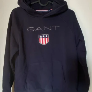 Mörkblå hoodie från GANT  - Snygg mörkblå hoodie från GANT med broderad logga och sköld på bröstet. Klassisk huva och stor magficka framtill. Perfekt för dig som gillar stilren och sportig look. Tillverkad i mjuk bomullsmix för extra komfort. Frakt tillkommer