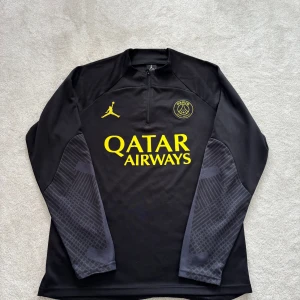 PSG x Jordan långärmad träningströja - Snygg svart långärmad PSG träningströja från Jordan med gul logga och Qatar Airways-tryck. Ärmarna har grått mönster och gul Paris-signatur. Halv dragkedja framtill och sportig passform, perfekt för fotbollsträning eller chill.