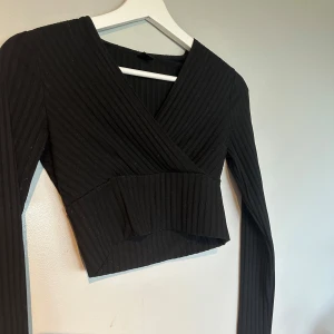 Svart omlott croppad ribbad topp - Snygg svart croppad topp med omlott-skärning och ribbad struktur. Långärmad modell som sitter tight. Super fin och jätte skönt material. 