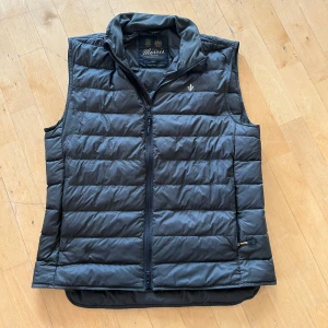 Mörkgrön dunväst från Morris - Snygg mörkgrön dunväst från Morris med quiltad design och hög krage i mjukt material. Västen har dragkedja framtill och en liten broderad logga på bröstet. Perfekt att slänga över en hoodie eller tröja när det är kyligt ute.