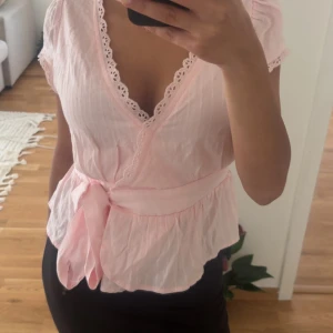 Söt rosa omlottblus med spets - Supersöt ljusrosa omlottblus med knyt i midjan och volang nertill. Blusen har korta ärmar och v-ringning med fina spetsdetaljer längs kanten. Perfekt för en romantisk look och passar till både jeans och kjol.
