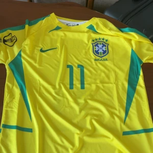 Brasilien fotbollströja - Gul Brasilien fotbollströja från Nike med gröna detaljer, nummer 11 och Ronaldinho på ryggen. Klassisk CBF-logga på bröstet och Nike Swoosh. Lätt och luftig polyester, perfekt för match eller träning.