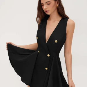 Svart kort klänning från Cider - Supersnygg svart kort klänning med djup v-ringning och guldiga knappar framtill. Klänningen är ärmlös och har en omlottlook med markerad midja och utställd kjol. Perfekt för dig som vill sticka ut med en chic och modern stil.