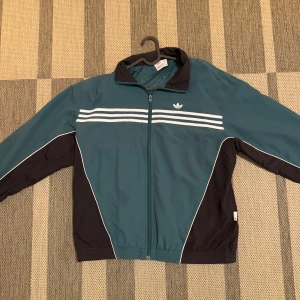 Adidas vindjacka i blått och svart - Snygg Adidas vindjacka i blått och svart med vita detaljer och klassiska ränder över bröstet och ryggen. Jackan har dragkedja, hög krage och coolt Adidas-tryck på ryggen. Perfekt för dig som gillar sportig och retro stil.