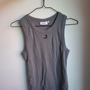 Grå ribbad linnetopp från Junkyard - Säljer en grå ribbad linnetopp från Junkyard i storlek XS. Toppen är ärmlös med rund halsringning och har en liten svart broderad logga framtill. Perfekt för en clean och enkel stil, lätt att matcha med jeans eller kjol. Toppen är inte använd men prislappen är borttagen. 