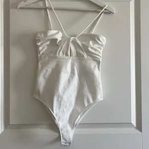 Vit bandeau topp från Zara - Supersnygg vit bandeau topp från Zara med tunna axelband och twistad detalj framtill. Toppen är ärmlös och har en lätt strukturerad yta i mjuk bomullsmix. Perfekt för varma dagar och enkel att styla med jeans eller kjol.