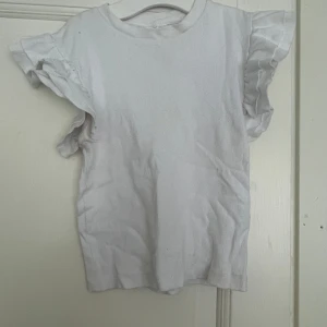 Vit volangtopp från Gina Tricot - Supersöt vit topp från Gina Tricot med ribbad struktur och volangdetaljer på ärmarna. Modellen är ärmlös med volanger som ger en extra touch. Perfekt för dig som gillar en enkel men ändå trendig look.