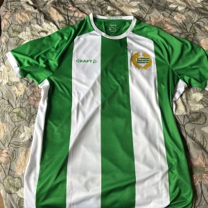 Hammarby fotbollströja från Craft - Snygg Hammarby fotbollströja från Craft med gröna och vita breda vertikala ränder. Tröjan har klubbmärket på bröstet och är tillverkad i ett lätt och ventilerande polyestermaterial. Klassisk passform med rund hals och korta ärmar.