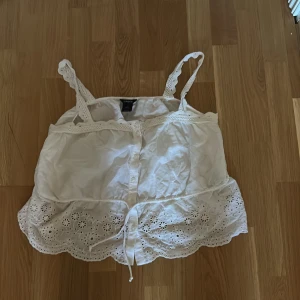 Vit broderad linnetopp från Lindex - Supersöt vit linnetopp från Lindex med broderade detaljer och spets på axelbanden. Toppen har knappar framtill, dragsko i midjan och en volangkant med blommiga broderier. Perfekt för varma dagar och ger en romantisk vibe.