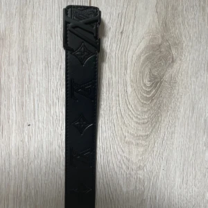 Svart Louis Vuitton skärp i skinn - Snyggt svart skärp från Louis Vuitton i skinn med präglade LV-loggor och monogramdetaljer längs hela bältet. Bältet har en mattsvart LV-spänne som ger en clean och lyxig vibe. Perfekt accessoar för att lyfta din outfit.