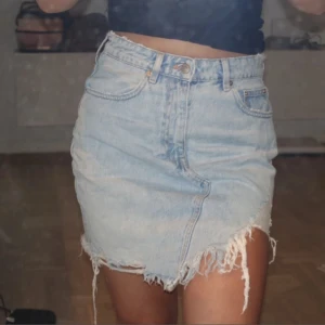 Ljusblå jeanskjol från Zara - Ljusblå jeanskjol från Zara med råa, fransiga kanter och asymmetrisk nederkant. Klassisk femficksmodell med bälteshällor och knappstängning. Perfekt för dig som gillar en avslappnad och trendig stil.