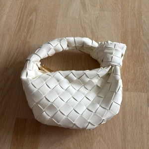 Vit flätad axelväska i skinn - Säljer en superfin vit axelväska med flätad design och knutdetalj på handtaget. Väskan är gjord i skinn och har en snygg guldfärgad dragkedja. Perfekt storlek för det viktigaste och ger en clean och trendig look.