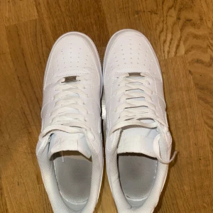Nike Air Force 1 helvita sneakers - Klassiska Nike Air Force 1 sneakers i helvitt skinn med vit sula och snörning. Modellen har perforerade detaljer på tån för extra ventilation och den ikoniska swoosh-loggan på sidorna. Perfekt för dig som gillar en clean och tidlös look. De är endast använda en gång och crease skydd tillkommer.