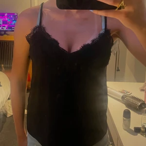 Svart linnetopp med spetsdetaljer - Säljer en svart linnetopp med tunna axelband och v-ringning. Toppen har fina spetsdetaljer längs urringningen som ger en extra touch. Perfekt festtröja eller bara en tröja till vardags!🩷
