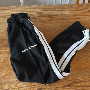 Svarta byxor med vita ränder från Palm Angels - Snygga svarta byxor från Palm Angels med vita ränder längs sidorna. Byxorna har en elastisk midja och dragkedja vid bensluten. Perfekta för en stilren och avslappnad look. 