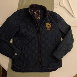 Mörkblå quiltad jacka från Morris - Säljer en stilren mörkblå quiltad jacka från Morris med broderad logga på bröstet. Jackan har dragkedja och knappar vid ärmsluten. Perfekt för våren
