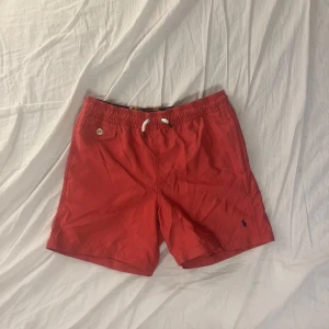 Röda badshorts från Polo Ralph Lauren - Snygga röda badshorts från Polo Ralph Lauren med elastisk midja och snörning. Shortsen har en liten broderad logga och en bakficka med knapp. Perfekta för sommarens stranddagar! Skriv vid minsta fundering! 👍