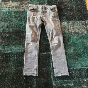 Grå jeans från Nudie Jeans - Snygga grå jeans från Nudie Jeans med klassisk femficksdesign och knappgylf. De har en straight passform och är perfekta för en avslappnad stil. Märkesdetaljer syns på knapparna och etiketten. Storlek W30 L32