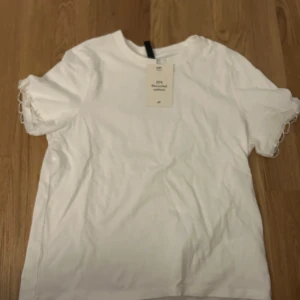 Vit t-shirt med pärldetaljer från H&M - Säljer en stilren vit t-shirt från H&M med unika pärldetaljer längs ärmarna. Gjord av 25% återvunnen bomull, perfekt för en hållbar garderob. Enkel och elegant design som passar till många tillfällen.