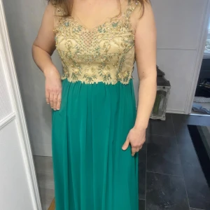 Grön långklänning med pärldetaljer från Radmela - Elegant grön långklänning från Radmela med en vacker överdel i beige dekorerad med pärlor och broderier. Klänningen har en ärmlös design och en flytande kjol som ger en fin silhuett. Perfekt för speciella tillfällen.