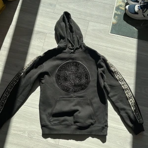 Svart hoodie från Glorious Gangsta - Säljer en svart hoodie från Glorious Gangsta med ett stort emblem på bröstet och mönstrade band längs ärmarna. Hoodien har en klassisk passform med känguruficka och justerbar huva. Perfekt för en stilren och avslappnad look.