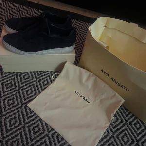 Mörk blåa sneakers från Axel Arigato - Snygga mörkblåasneakers från Axel Arigato med en stilren design. Skorna har en vit sula som kontrasterar snyggt mot det svarta materialet. Perfekta för en trendig och avslappnad look.