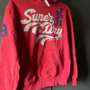 Röd hoodie från Superdry - Säljer en röd hoodie från Superdry med stor vit logga på framsidan och blå detaljer. Tröjan har en klassisk känguruficka och justerbar huva med snören. Perfekt för en avslappnad stil.
